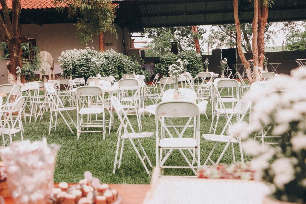 An outdoor wedding venue setup with white folding chairs on green grass and decorative plants.